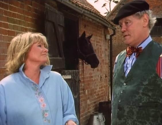 Tinker in beret and bow tie talking to a woman outside a brick stable, a horse looking out over the door behind them