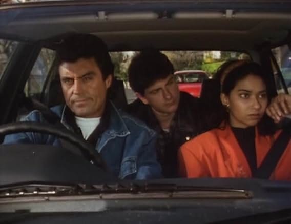 Lovejoy at the wheel of Susan's car in a denim jacket, Eric leaning forward from the back seat, Khadija beside him in an orange jacket, all three looking grim